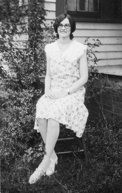 Luna Early. She sits with her legs crossed. She is wearing glasses and a dress and she has medium hair. She is smiling.