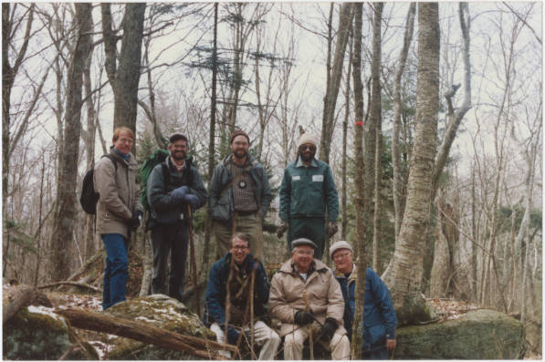 Group of 7 hikers in a wooded area. They are dressed warmly and are all smiling.