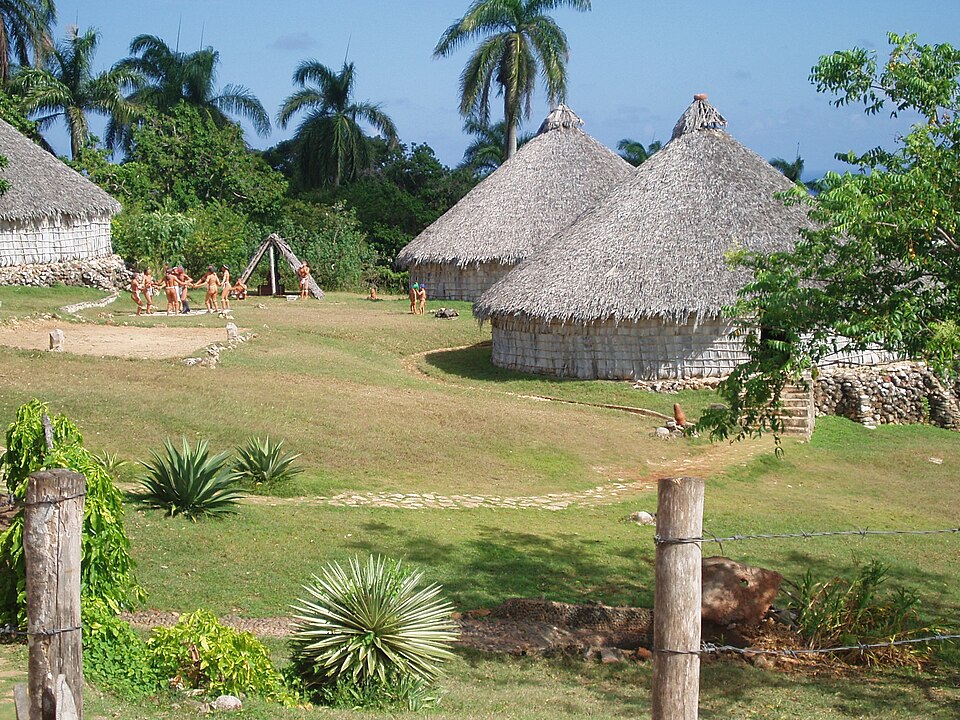 A small village of huts sits in a tropical landscape. People gather in the background.
