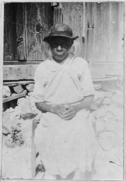 Lila Nichols. She is seated in a chair beside a wooden structure. She is wearing a simple dress and leather hat. Her hands are clasped.