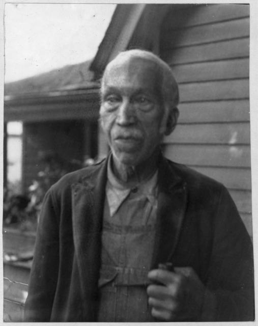 Bost. He is pictured wearing a coat and overalls. He is older, with short white hair and a thin mustache. A small house sits behind him.