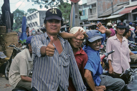 Three smiling men wave and gesture in a Chinatown market at Ho Chi Minh City
